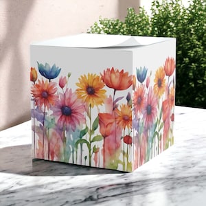 Puede incluir: Un bloc de notas cuadrado blanco con un diseño de acuarela de flores coloridas. Las flores son en tonos de rosa, rojo, amarillo, azul y verde.