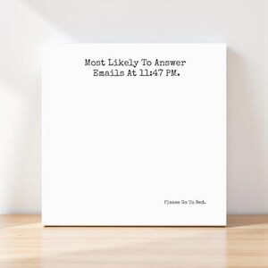 May include: A white square canvas with the text "Most Likely To Answer Emails At 11:47 PM." in a typewriter font. Below, the text "Please Go To Bed." is printed in the same font. The canvas is on a wooden surface.