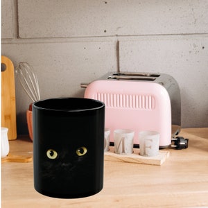 May include: Black ceramic mug with a black cat face design. The cat's eyes are yellow. The mug is on a wooden surface next to a pink toaster and white cups.