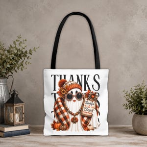 May include: A white tote bag with black handles featuring a graphic of a ghost wearing a plaid scarf, a beanie, and round sunglasses. The ghost holds a sign that reads Thankful & Blessed, surrounded by autumn pumpkins and fall foliage.
