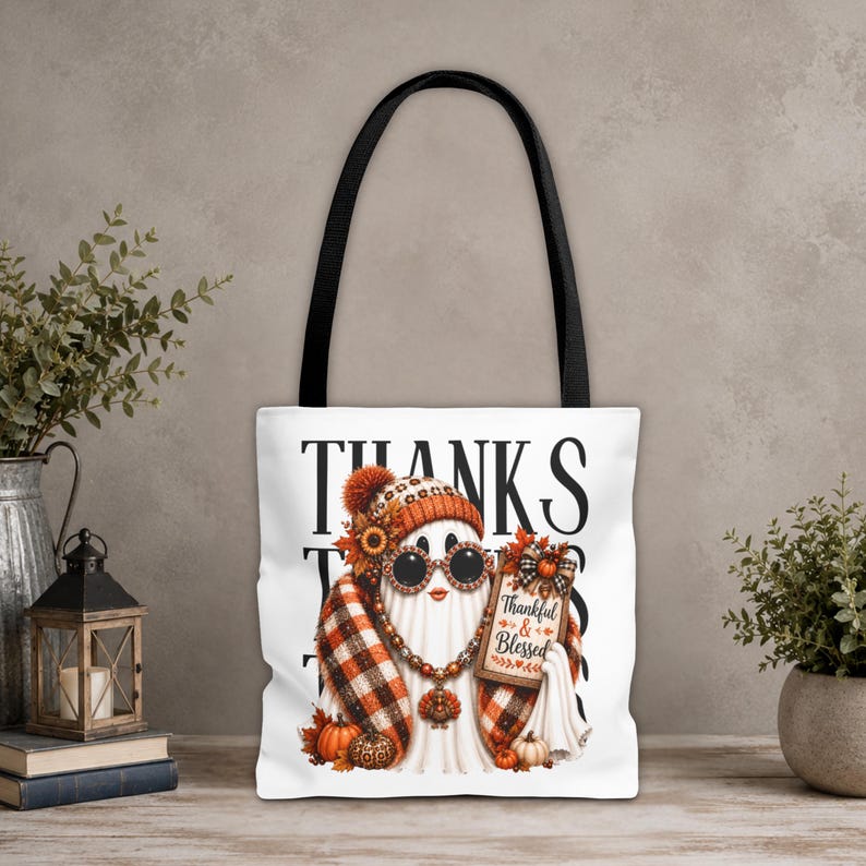 May include: A white tote bag with black handles featuring a graphic of a ghost wearing a plaid scarf, a beanie, and round sunglasses. The ghost holds a sign that reads Thankful & Blessed, surrounded by autumn pumpkins and fall foliage.