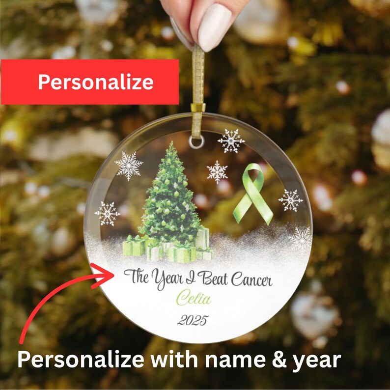 May include: Clear, round Christmas ornament with a green ribbon, Christmas tree, and snowflakes. The ornament has the text "The Year I Beat Cancer Celia 2025". The ornament is held by a gold ribbon.