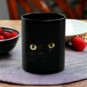 May include: Black ceramic mug with a black cat peeking out from behind the rim. The cat has bright yellow eyes.