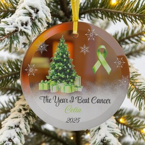 May include: Round glass Christmas ornament with a gold ribbon. The ornament features a green Christmas tree, gifts, snowflakes, and a green ribbon. Text reads "The Year I Beat Cancer Celia 2025."