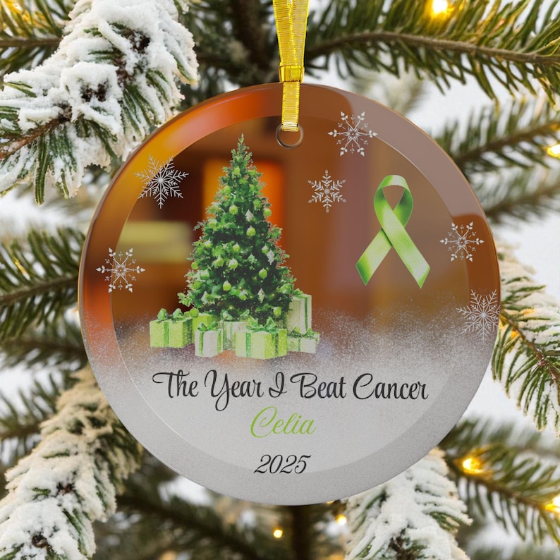 May include: Round glass Christmas ornament with a gold ribbon. The ornament features a green Christmas tree, gifts, snowflakes, and a green ribbon. Text reads "The Year I Beat Cancer Celia 2025."