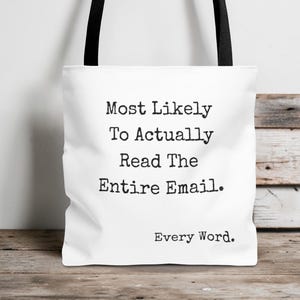 Op de afbeelding: Witte draagtas met zwarte handvatten. De tas heeft de tekst "Most Likely To Actually Read The Entire Email. Every Word." in een lettertype in typemachinestijl. De tas staat op een houten oppervlak.