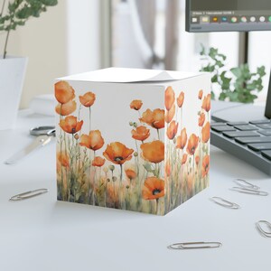 May include: A white cube-shaped notepad with a watercolor design of orange poppies on a white background. The notepad is on a white surface.