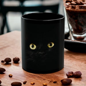 May include: Black ceramic mug with a black cat peeking out from the darkness, its yellow eyes staring directly at the viewer.