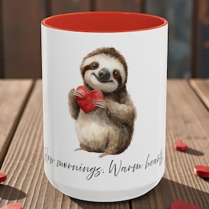 May include: White ceramic mug with a red interior featuring a watercolor illustration of a smiling sloth holding a red heart. The mug has the text "Slow mornings. Warm hearts."