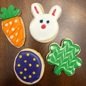 May include: Four decorated sugar cookies, one shaped like a carrot, one like a bunny, one like a shamrock, and one round with blue and yellow icing.