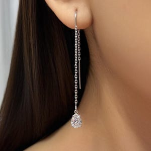 Op de afbeelding: Zilveren oorbel met een peervormige, heldere edelsteen. De oorbel hangt aan het oor met een fijne ketting. De edelsteen is facetgeslepen en vangt het licht.