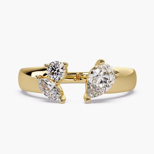 Può includere: Un anello d'oro con tre pietre preziose trasparenti: un taglio rotondo, un taglio a marquise e un taglio a goccia. L'anello ha un design a gambo diviso, con le pietre preziose raggruppate nella parte superiore.