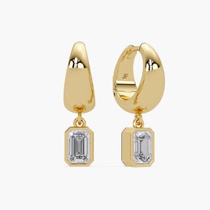 May include: A pair of gold-coloured hoop earrings with a rectangular, clear gemstone pendant. The earrings have a polished finish and a hinged closure. The gemstones are set in a bezel setting.