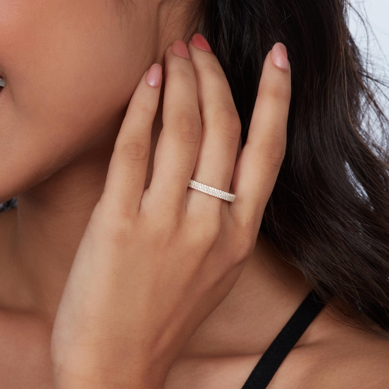 以下が含まれることがあります： 小さな透明な宝石で覆われたバンドが付いた、金色のリング。 リングは指に着用されており、画像には人の手と腕が見えます。 このリングは結婚指輪またはエタニティリングのようです。