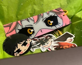 May include: A collection of anime-themed accessories, including a pink and gray bookmark with a character's eyes, a black round pin with a character's face, and two character stickers, all resting on green tissue paper.