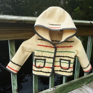 May include: Cream-colored hooded jacket with horizontal stripes in red, green, and black. The jacket has two front pockets and is trimmed with black and white patterned fabric. The jacket is displayed on a wooden railing.