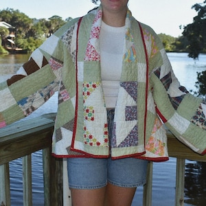 May include: A quilted jacket featuring a patchwork design with various fabric patterns in shades of green, white, and floral prints. The garment has wide sleeves, an open front, and is finished with a thin red trim along the edges and hemline.