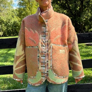 May include: A textured, long-sleeved jacket in shades of peach, orange, and light green with a large leaf-like pattern. The jacket features a high collar, a central button placket with a brown and white plaid trim, and two large front pockets.