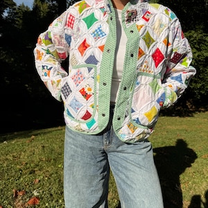 May include: A quilted jacket constructed from a patchwork of multi-colored fabric squares in various patterns, featuring a light green patterned trim along the collar, front opening, and two front pockets, secured with silver-toned metal snap buttons.