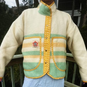 May include: Cream-coloured jacket with a high collar and button closure. The jacket features a green and yellow striped design across the chest and pockets, with a small emblem on the left pocket. The collar and button placket are accented with a yellow floral pattern.