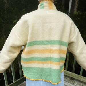 May include: Cream wool jacket with yellow and green horizontal stripes. The jacket has a high collar and long sleeves. The bottom and sleeve edges are trimmed with yellow decorative trim. The jacket is worn over a light blue garment.
