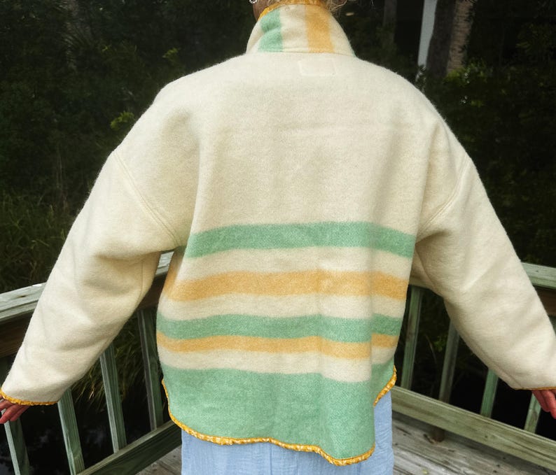May include: Cream wool jacket with yellow and green horizontal stripes. The jacket has a high collar and long sleeves. The bottom and sleeve edges are trimmed with yellow decorative trim. The jacket is worn over a light blue garment.