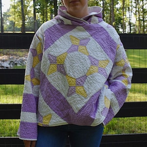 May include: A quilted hoodie in a patchwork design. The hoodie features a pattern of white, lavender, and yellow starbursts and stripes. The garment is worn over blue jeans. The background includes a black fence and green foliage.