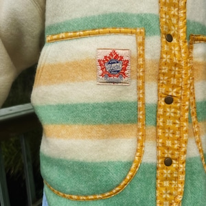 May include: Close-up of a vintage wool jacket with cream, green, and yellow horizontal stripes. The jacket has a patch pocket and is trimmed with yellow fabric. The label reads "Ayers Wool Canadian".