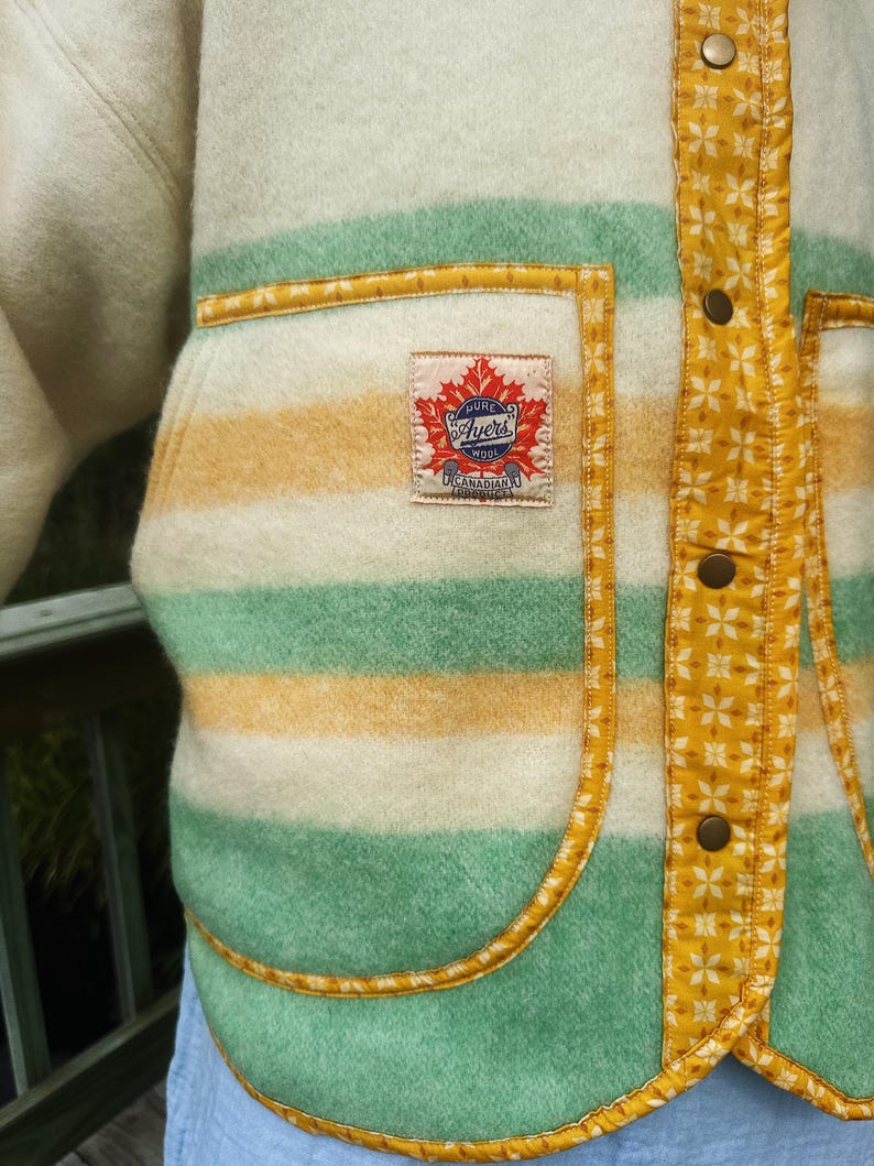 May include: Close-up of a vintage wool jacket with cream, green, and yellow horizontal stripes. The jacket has a patch pocket and is trimmed with yellow fabric. The label reads "Ayers Wool Canadian".