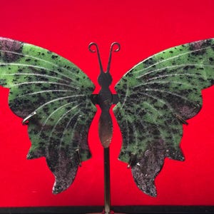 Peut inclure: Un objet décoratif en forme de papillon taillé dans de la zoïsite verte avec des inclusions noires et rouge foncé. Les ailes en pierre sont fixées sur un support métallique vertical fin, doté d'un corps central avec deux antennes courbées.