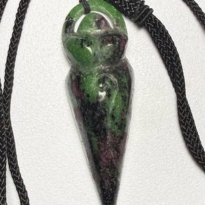 May include: A pendant necklace featuring a carved gemstone pendant with a teardrop shape. The pendant is green with dark red and black inclusions, suspended from a black braided cord. The pendant is approximately 2 inches long.