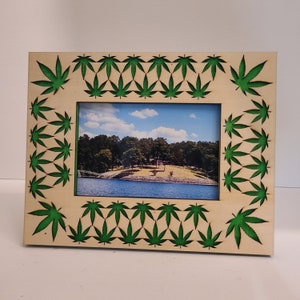 Może przedstawiać: Drewniana ramka na zdjęcia z zielonym wzorem liści marihuany. Ramka jest pusta i gotowa do włożenia zdjęcia.