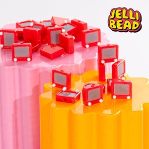 May include: A collection of red, miniature TV-shaped candies with gray screens and white accents. The candies are arranged on pink and yellow, wavy-edged platforms. The image includes the text "JELLI BEAD" in a colorful, bubble-letter style.