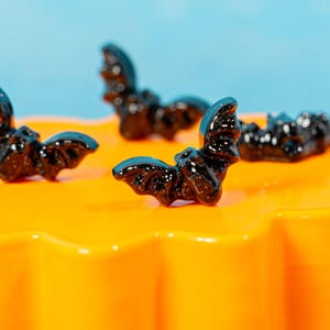 May include: Three small, black, glittery bat-shaped figurines are positioned on a bright orange surface. Each bat features detailed wings and a small head, with a glossy finish that reflects light, creating a distinct contrast against the orange base.