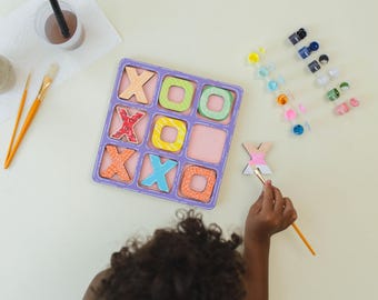 Può includere: Un bambino dipinge una X di legno con un pennello. La X fa parte di un gioco di tris con pezzi di legno colorati. Il tabellone di gioco è viola e ha uno sfondo rosa. Ci sono barattoli di vernice sullo sfondo.