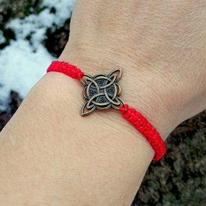 以下が含まれることがあります： 赤い紐のブレスレットに、ブロンズ製のケルト結び目のチャームが付いています。ブレスレットは人の手首に結ばれています。