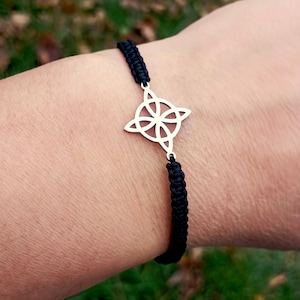 May include: A black woven bracelet featuring a silver-colored Celtic knot charm. The charm is a four-pointed design with an interwoven pattern. The bracelet is worn on a wrist, with the background out of focus.