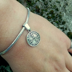 Puede incluir: Una pulsera de plata con un colgante redondo que presenta un diseño de cruz. El colgante tiene una superficie texturizada y detallada.