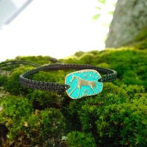 May include: A black macrame bracelet featuring a rectangular, turquoise-colored charm with a bronze-colored horse silhouette. The charm has a distressed, vintage appearance. The bracelet is displayed on a bed of green moss.