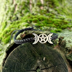 May include: A black braided bracelet with a silver charm featuring a triple moon and pentagram symbol. The bracelet is on a mossy tree trunk.