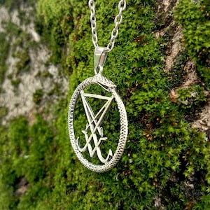 以下が含まれることがあります： 銀色のネックレスに、五芒星に巻き付いた銀色のヘビのペンダントが飾られています。ペンダントの中にはシンボルがあります。ネックレスは緑色の苔を背景にしています。