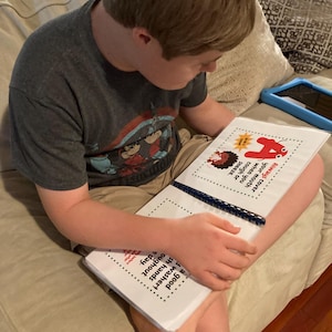 Op de afbeelding: Een jong persoon zit op een bank en leest een boek met een rood nummer vier op de omslag. Het boek heeft de tekst "Bedek altijd je mond wanneer je hoest of niest. Een goede wassen! Was je handen de hele dag door."