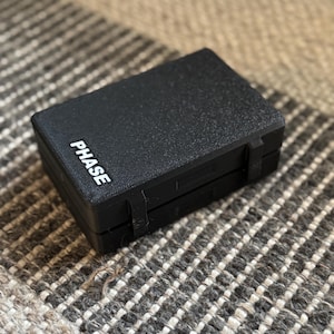 May include: A black, rectangular box with the word "PHASE" printed in white on the side. The box has a textured surface and appears to be made of plastic.