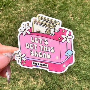 May include: Pink toaster sticker with a glittery finish. The toaster has dollar bills inside and the text "Let's Get This Bread" on the front. The text "Hot & Fresh" is printed below the toaster.