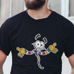 May include: Black t-shirt featuring a cartoon character with a white face, large eyes, and yellow hands. The character's body is a colorful, segmented design. The shirt has rolled-up sleeves and is worn by a person wearing sunglasses and a black cap.