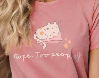 Puede incluir: Camiseta rosa claro con una ilustración de un gato durmiendo y el texto "Nope. Too peopley." El gato es rosa claro con manchas naranjas, descansando sobre una almohada rosa. También se incluyen pequeñas flores amarillas.