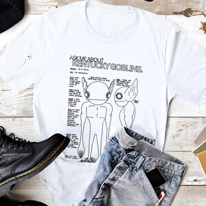 K&ouml;nnte beinhalten: Wei&szlig;es T-Shirt mit dem Text "ASK ME ABOUT KENTUCKY GOBLINS" und einer detaillierten Illustration von zwei goblinartigen Kreaturen. Das Shirt wird mit blauen Jeans, einer schwarzen M&uuml;tze und schwarzen Stiefeln kombiniert. Auch wei&szlig;e Ohrh&ouml;rer sind zu sehen.