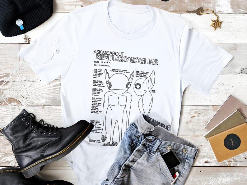K&ouml;nnte beinhalten: Wei&szlig;es T-Shirt mit dem Text "ASK ME ABOUT KENTUCKY GOBLINS" und einer detaillierten Illustration von zwei goblinartigen Kreaturen. Das Shirt wird mit blauen Jeans, einer schwarzen M&uuml;tze und schwarzen Stiefeln kombiniert. Auch wei&szlig;e Ohrh&ouml;rer sind zu sehen.