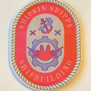 May include: A white oval patch with a beige rope border. The center features a red background with the text "SHIPKIN SHIPPA SHIPBUILDING". Inside the text is a purple gear with a red bunny inside. Above the gear is a red anchor with a white cross on each side.