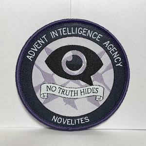 Puede incluir: Parche redondo con el texto "ADVENT INTELLIGENCE AGENCY" en letras blancas sobre un borde morado oscuro. El parche presenta un gráfico de ojo estilizado y las palabras "NO TRUTH HIDES" en una pancarta. La palabra "NOVELITES" está en la parte inferior.
